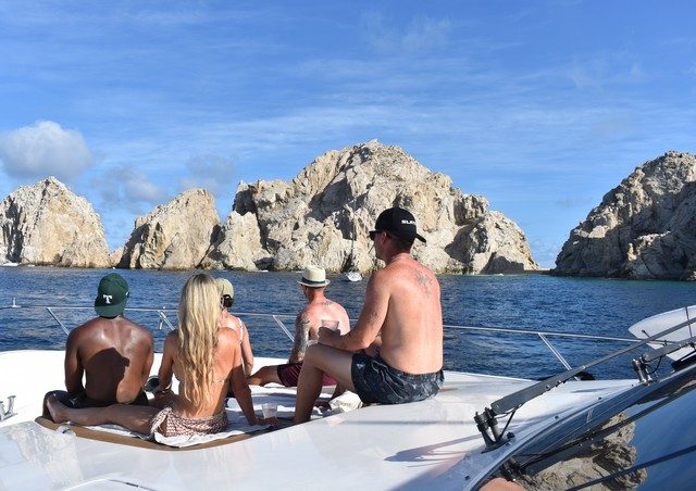 Friends relaxing on the bow of a yacht with El Arco ahead