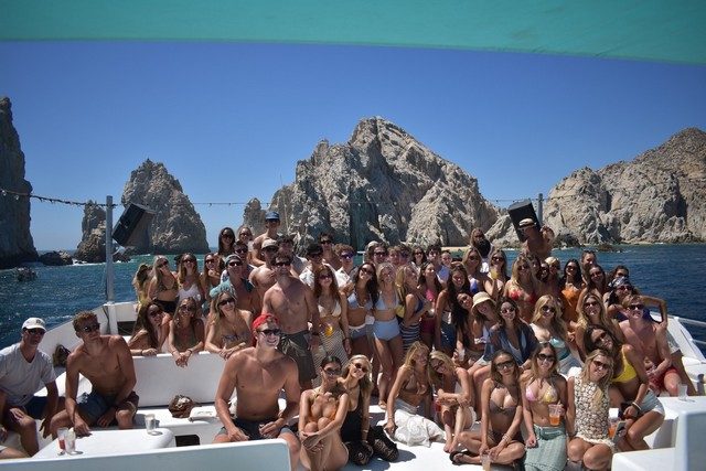 Large group celebrating on a boat deck with El Arco behind them