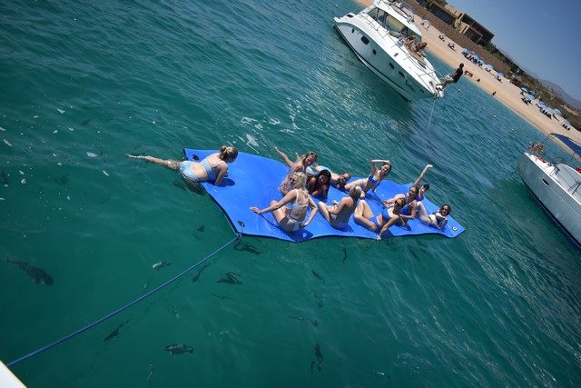 Group floating on a water mat beside an anchored yacht